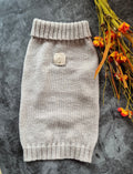 A light beige knit pet sweater with ribbed cuffs and hem, laid flat on a dark gray textured surface next to dried orange flowers.