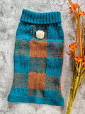 A hand-knit pet sweater in teal and rust-orange plaid, laid flat on a textured gray surface next to dried orange flowers.