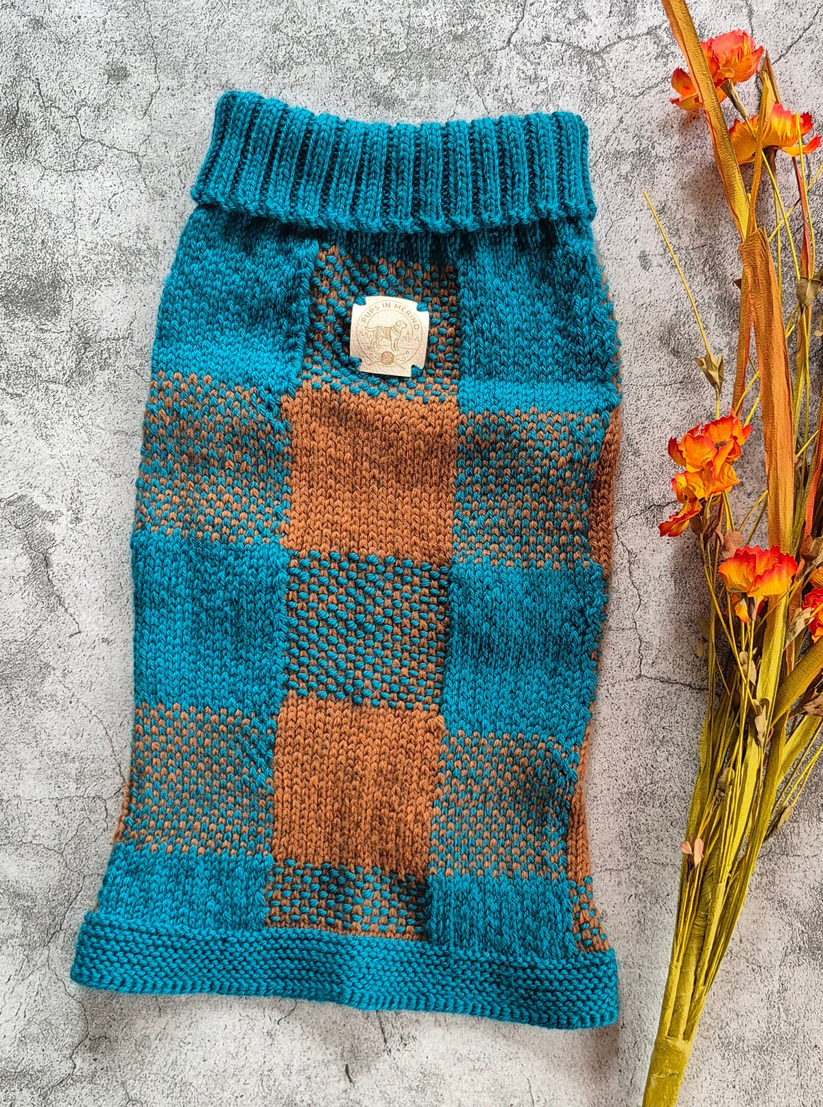 A hand-knit pet sweater in teal and rust-orange plaid, laid flat on a textured gray surface next to dried orange flowers.