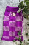 A purple and white checkered knit sweater vest laid flat on a textured gray surface, styled with green trailing houseplant vines.