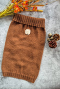 A brown knit pet sweater laid flat on a gray textured surface, styled with dried orange flowers and pinecones. The sweater has a ribbed collar, cuffs, and hem, and a small square label patch near the upper center.