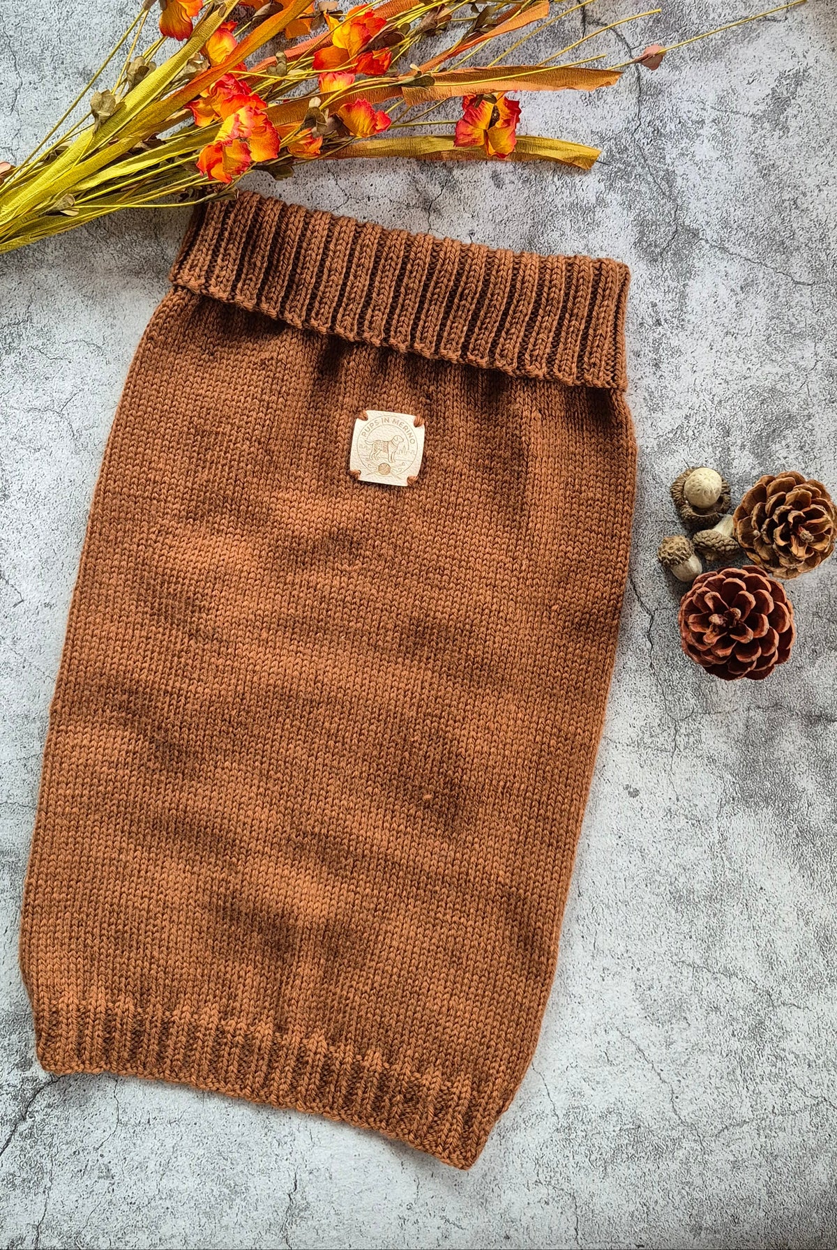 A brown knit pet sweater laid flat on a gray textured surface, styled with dried orange flowers and pinecones. The sweater has a ribbed collar, cuffs, and hem, and a small square label patch near the upper center.