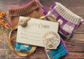 A gift card with a ribbon loop and decorative seal reading 'Gift Card' and 'Pups in Merino' lies on top of folded knit dog sweaters in brown, purple, cream, and teal, arranged on a rustic wooden surface with dried orange flowers.